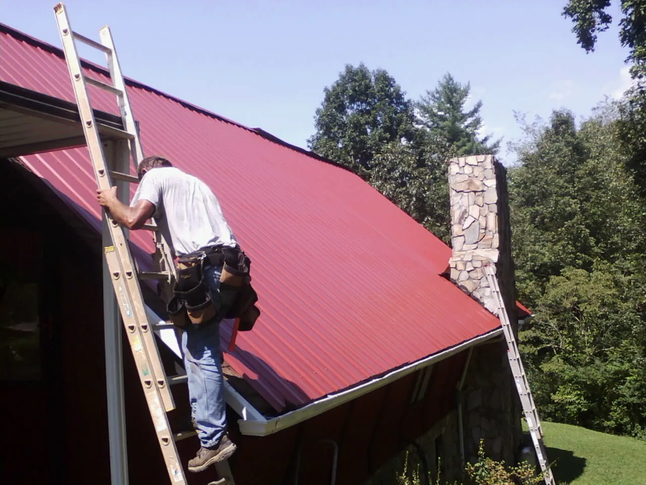 Roofer on a ladder beside a red metal roof and stone chimney — Soffit & Fascia Repair by Roofing Brothers in Bakerstown